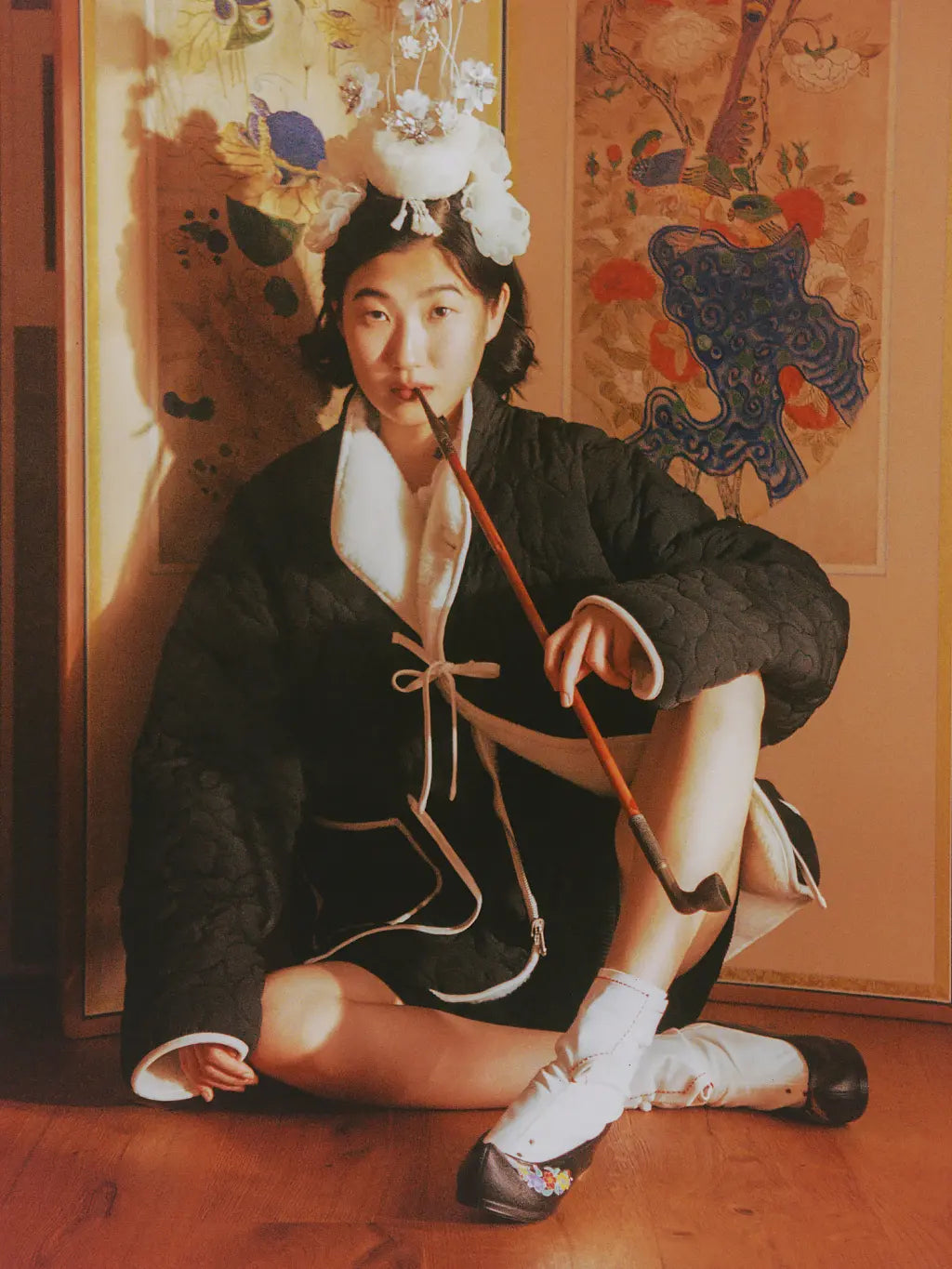 A person wearing a Sundae School Reversible Flower Quilted Fleece and a decorative white floral headpiece sits on a wooden floor indoors, holding a long brown object. The background shows warm-toned wall panels with painted motifs.