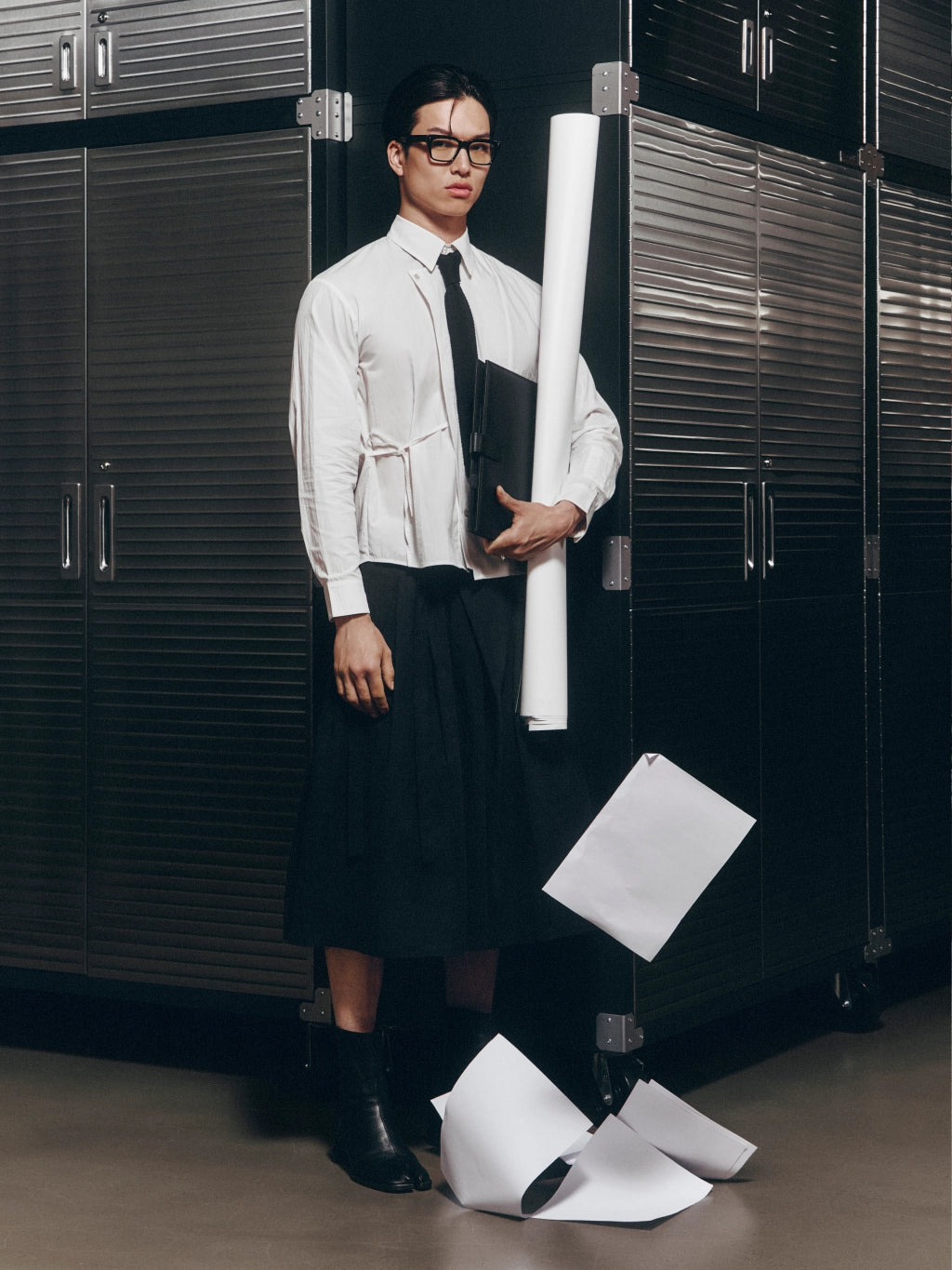 A man wearing a white long-sleeve shirt with a tie and a long black skirt stands in front of dark metal cabinets, holding a rolled sheet of paper. Several loose sheets are scattered on the floor around him.