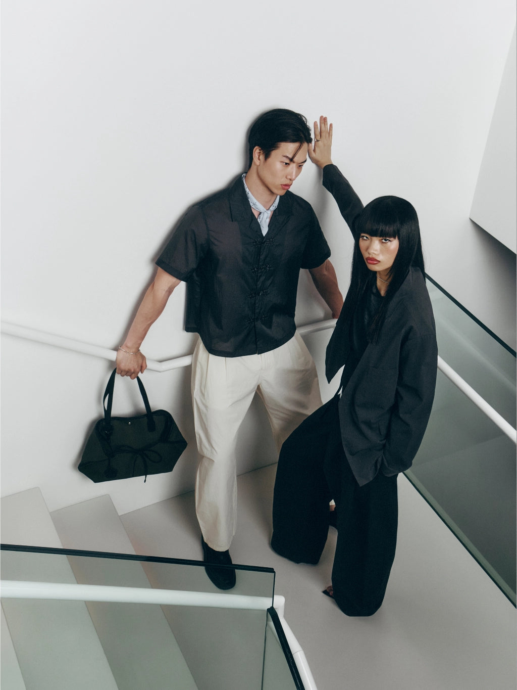 A man and a woman pose on a modern white staircase. The man wears a black short-sleeve button-up shirt, cream trousers, and holds a black handbag.