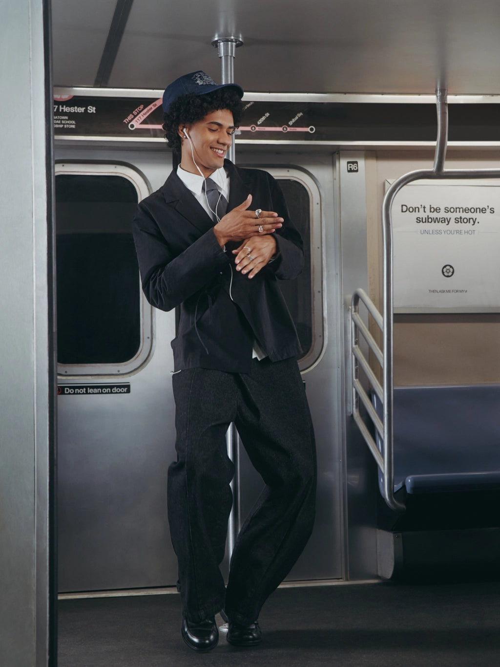 A man in a dark navy blue uniform with a matching cap stands inside a subway train car, smiling and posing. The uniform resembles a conductor's outfit, with a dark jacket, trousers, and a tie.