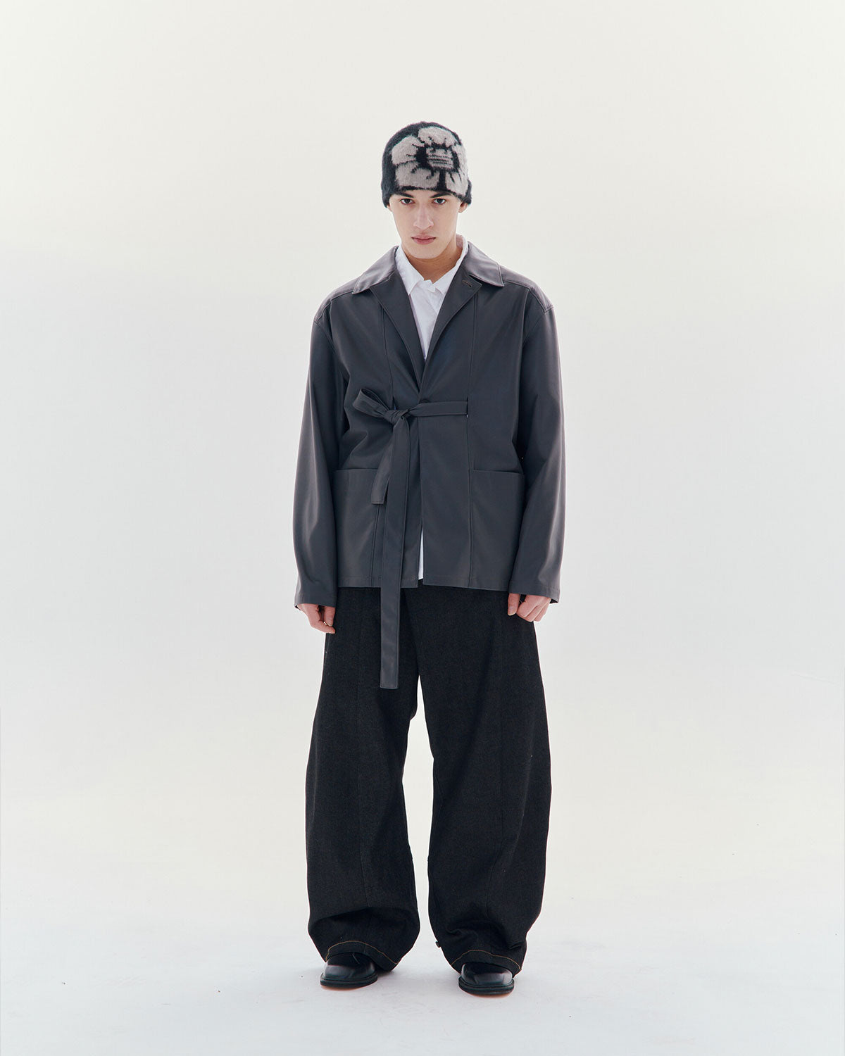 Against a plain white background, the person wears Sundae School's Black Ddul Beanie with a gray belted jacket, white shirt, wide-leg dark pants, and black shoes—a minimalist, modern look.