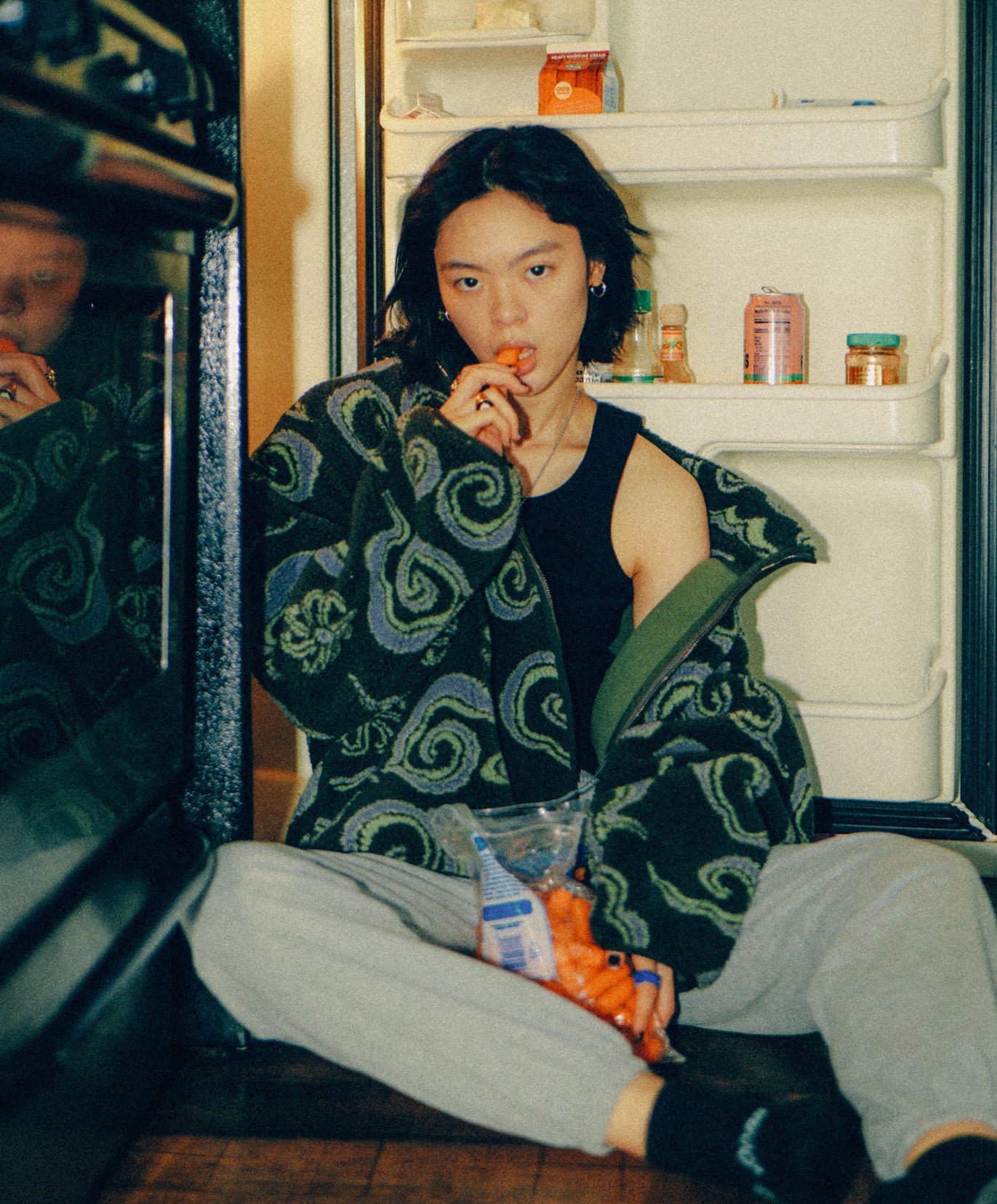 A young woman with shoulder-length dark hair sits in front of an open refrigerator, wearing a dark tank top and an oversized Sundae School Kloud Fleece jacket with spiral patterns. She is eating baby carrots while looking at the camera. The refrigerator contains various jars and containers on the shelves.