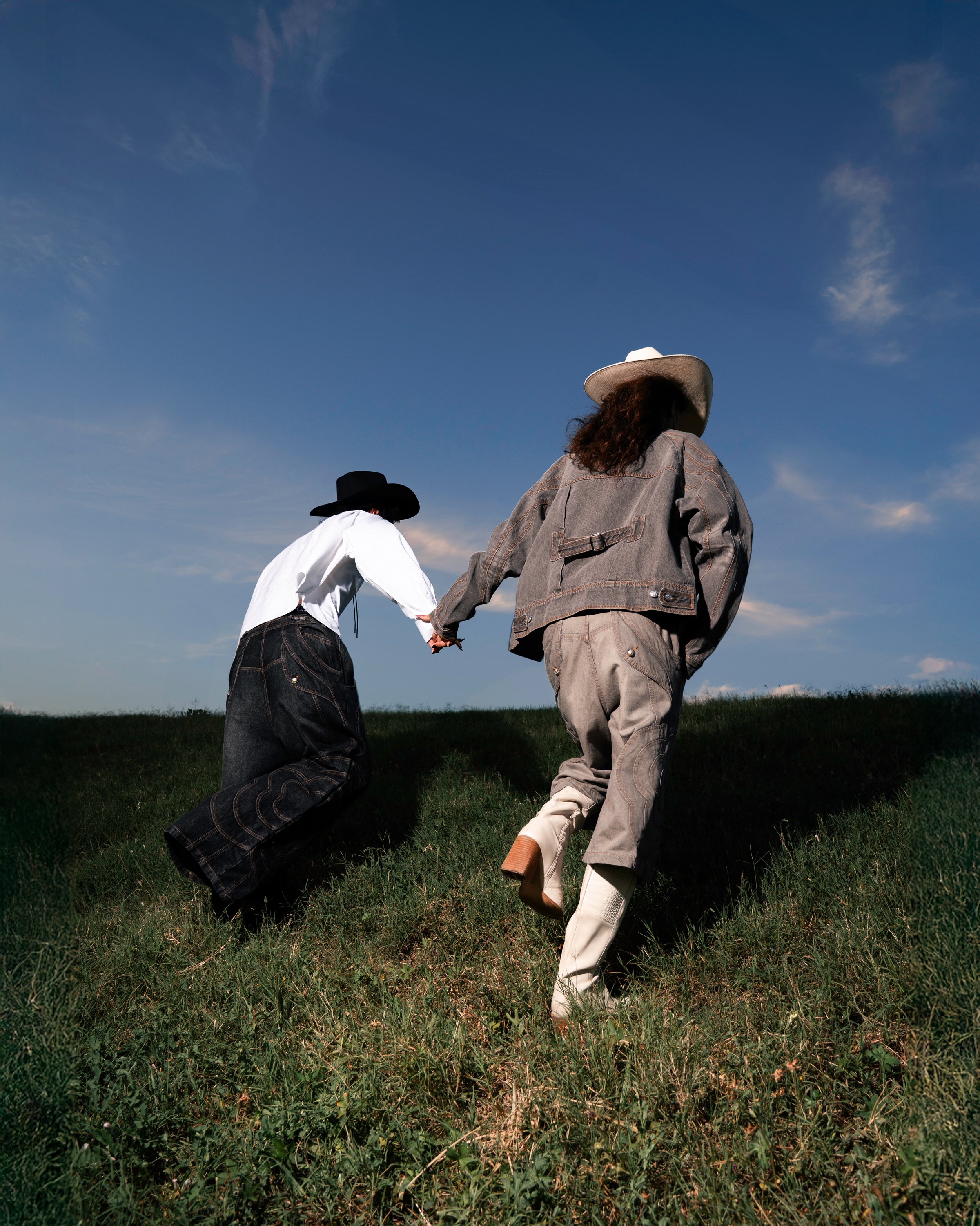 Two people in wide-brimmed hats and western-style clothing—one wearing Sundae School's BLACK MOCHI PANELED PANTS (x JOONIVERSE)—walk hand in hand up a grassy hill under a clear blue sky, photographed from behind.