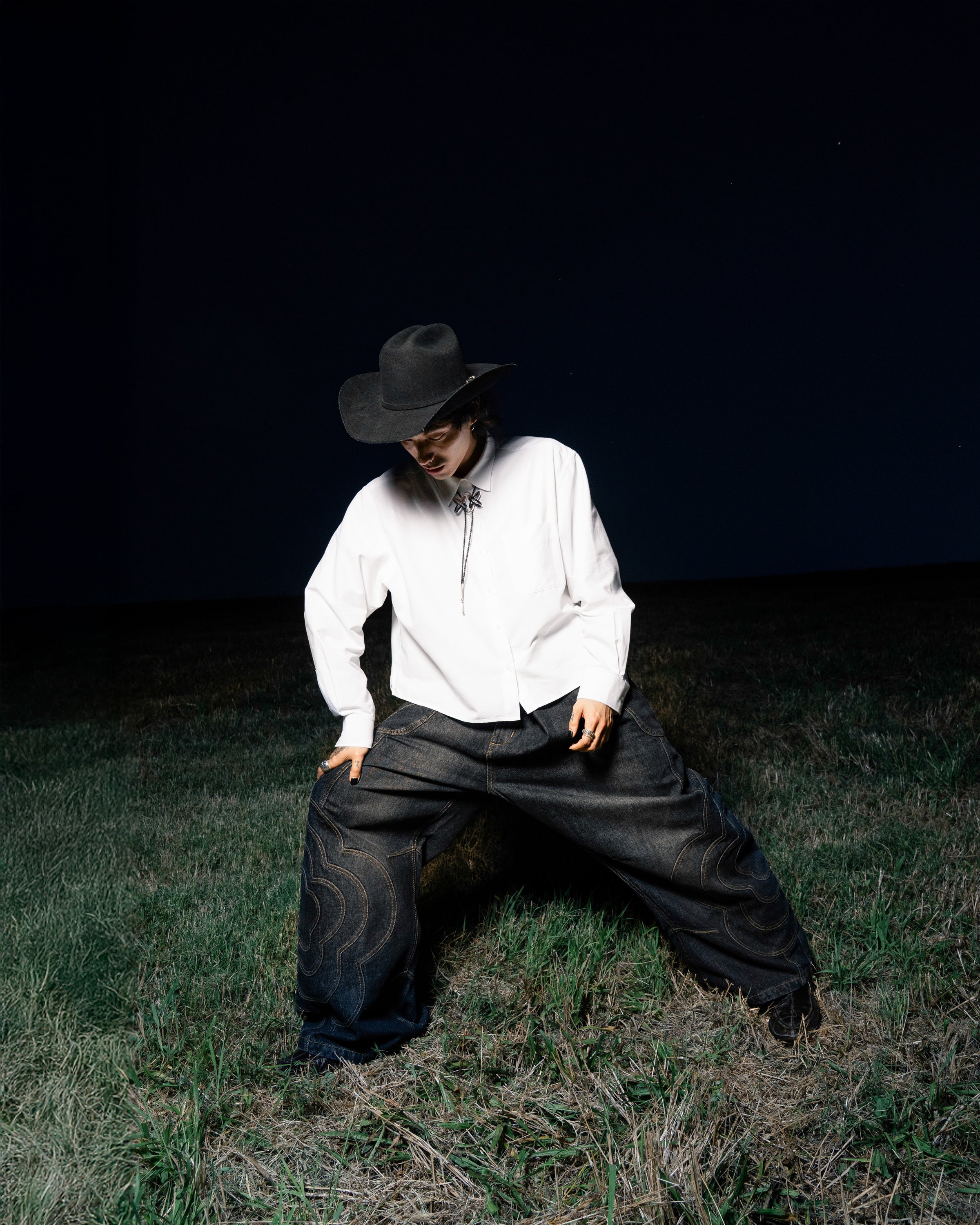A person in a wide-brimmed black hat, sunglasses, white shirt, and Sundae School BLACK MOCHI PANELED PANTS (x JOONIVERSE) stands on grass at night with legs spread apart, looking down.