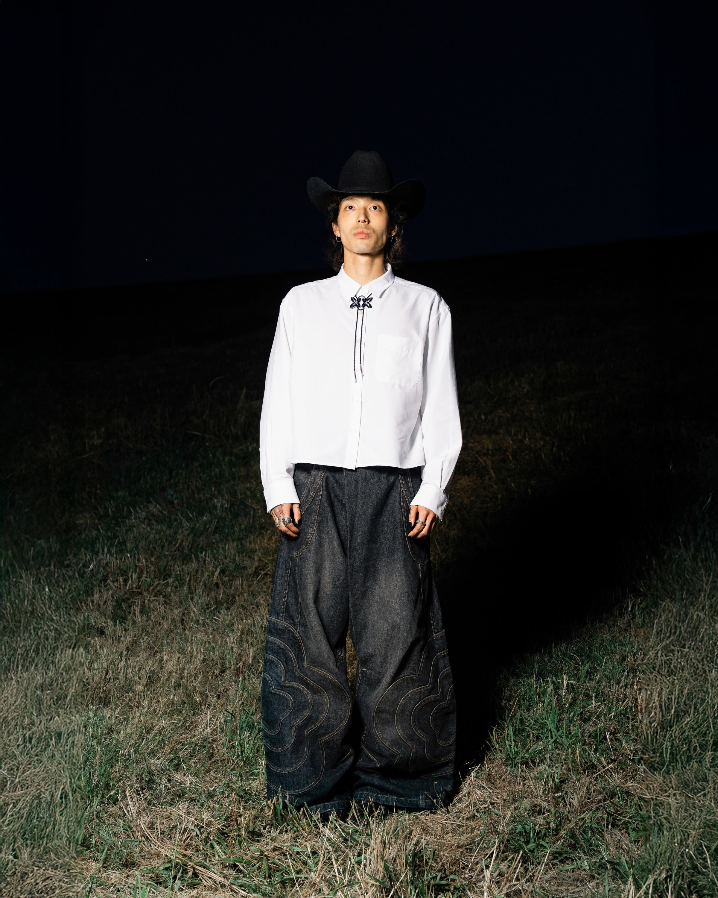 A person in a white shirt, BLACK MOCHI PANELED PANTS (x JOONIVERSE) from Sundae School, and a black cowboy hat stands in a grassy field at night under a spotlight.