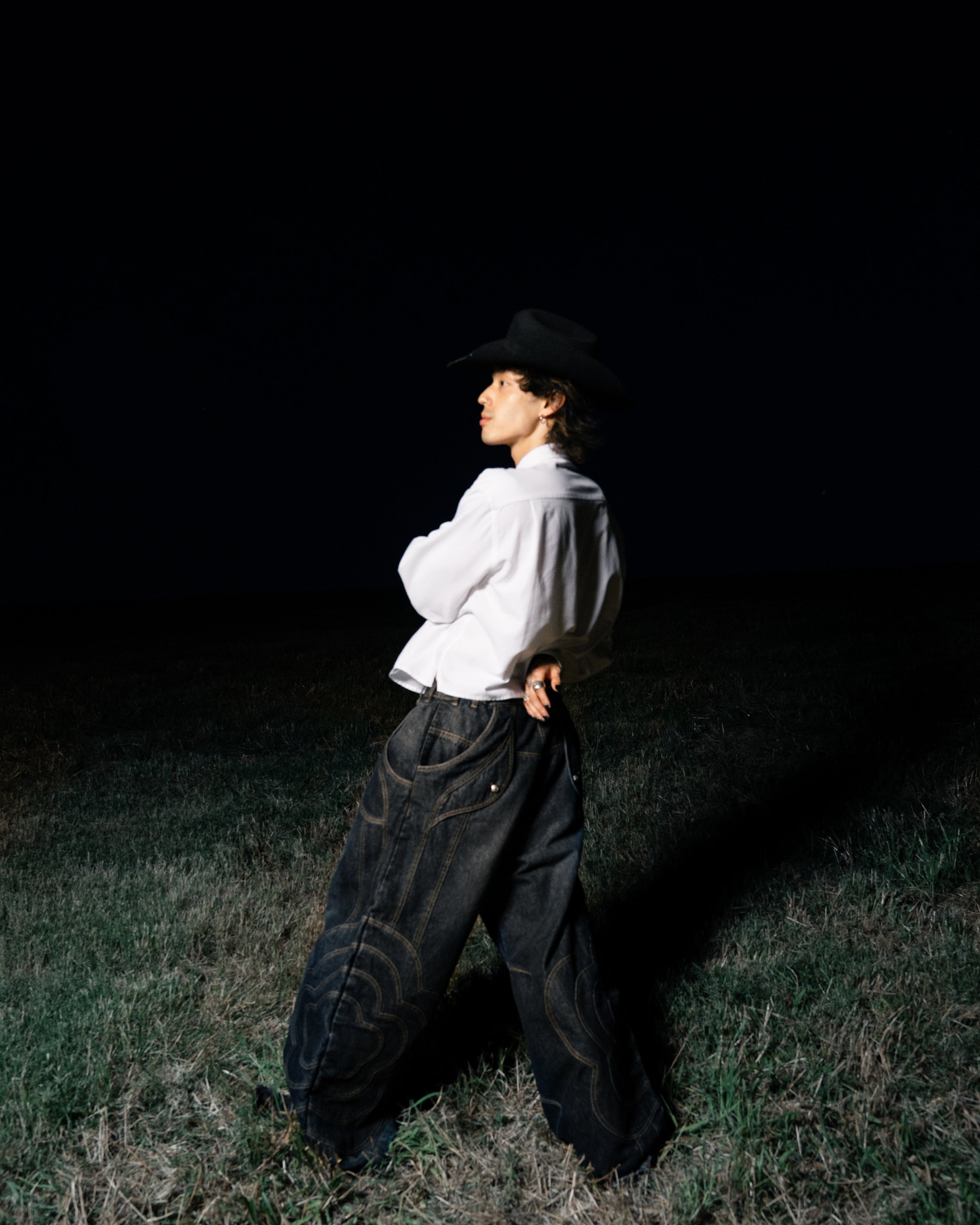 At night, a person wearing a wide-brimmed black hat, white shirt, and Sundae School BLACK MOCHI PANELED PANTS (x JOONIVERSE) stands on grass facing left with arms crossed behind their back against a dark background.