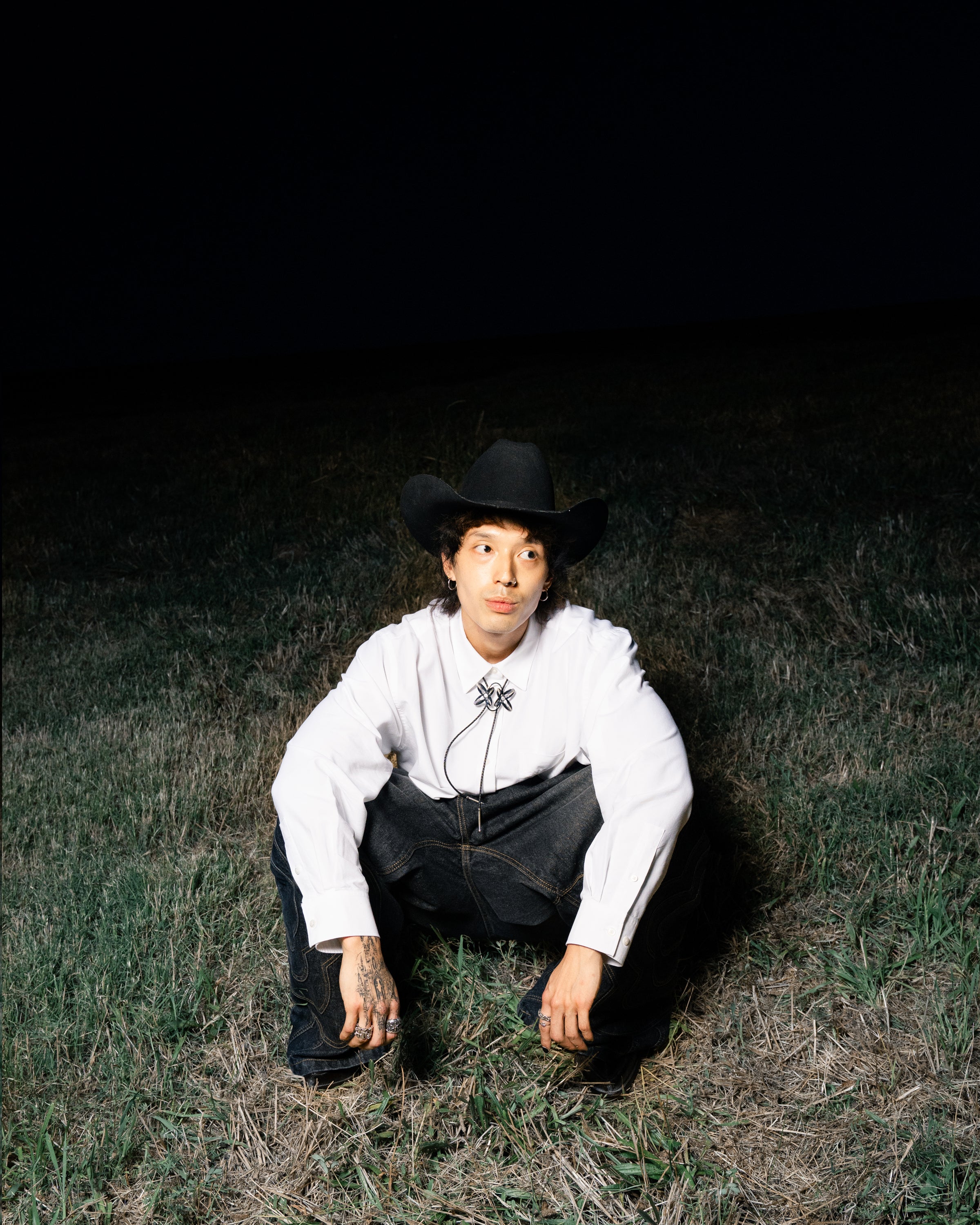 A person in a black cowboy hat, white shirt, bolo tie, and Sundae School BLACK MOCHI PANELED PANTS (x JOONIVERSE) squats on grass at night. The illuminated subject stands out against the dark background.