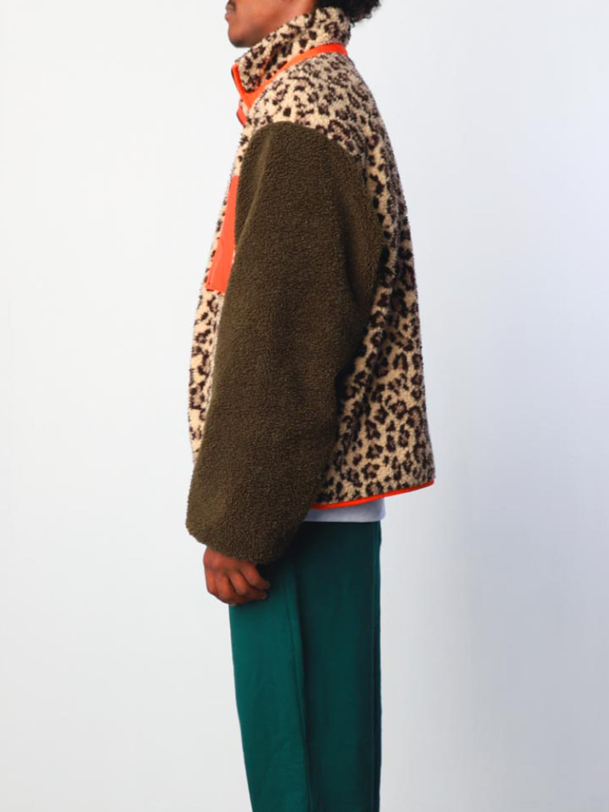 A person stands in profile against a plain light background, wearing Sundae School's Tiger Mom Fleece—a sherpa jacket in leopard print and olive green with an orange accent—and teal pants.