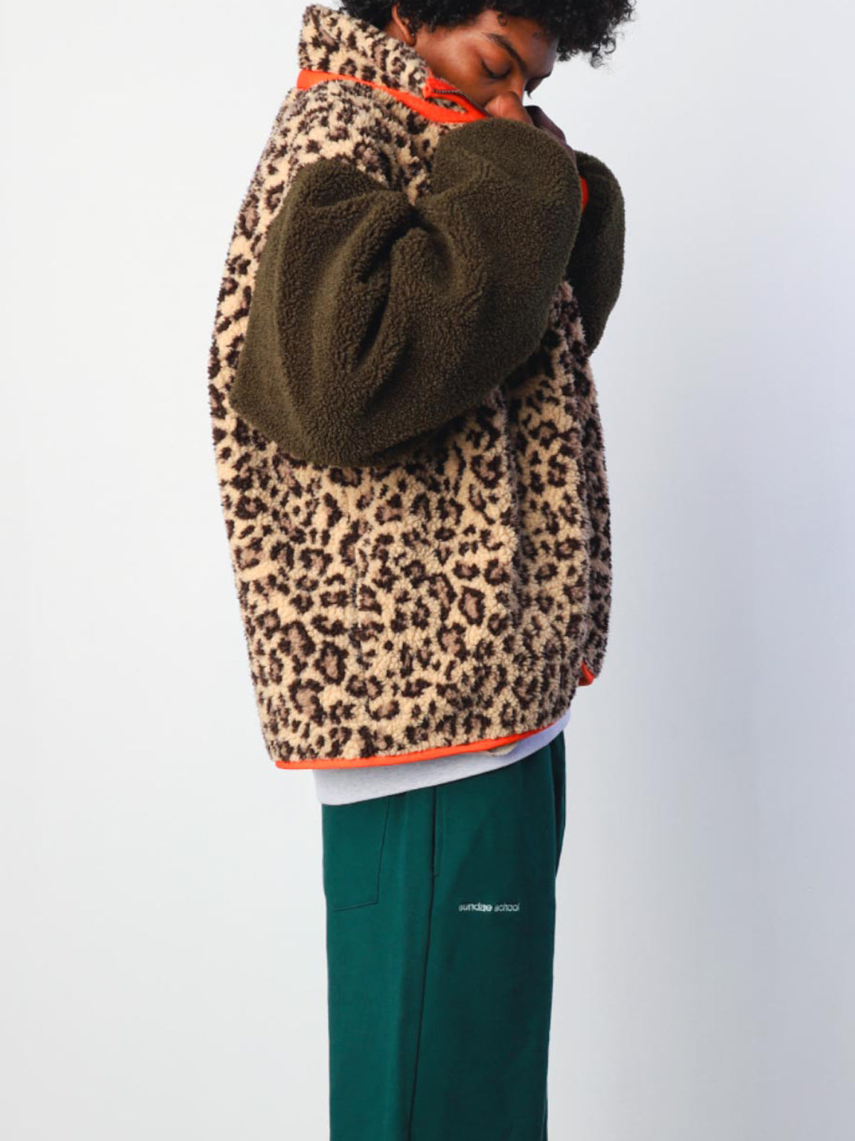 A person wears the Sundae School Tiger Mom Fleece, featuring brown and black sherpa with dark green sleeves, paired with green pants. They stand sideways and look down against a plain white background.