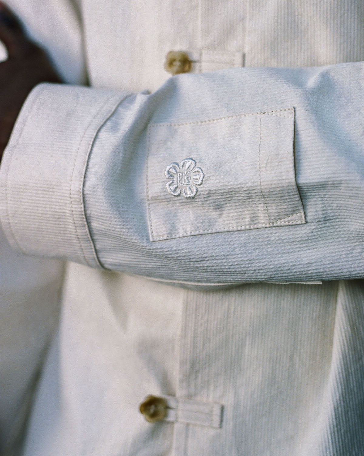 A close-up of a person wearing the Sundae School IVORY CHINA COLLAR KNOT SHACKET, a light corded cotton twill shirt with beige buttons and a sleeve pocket featuring an embroidered white flower.