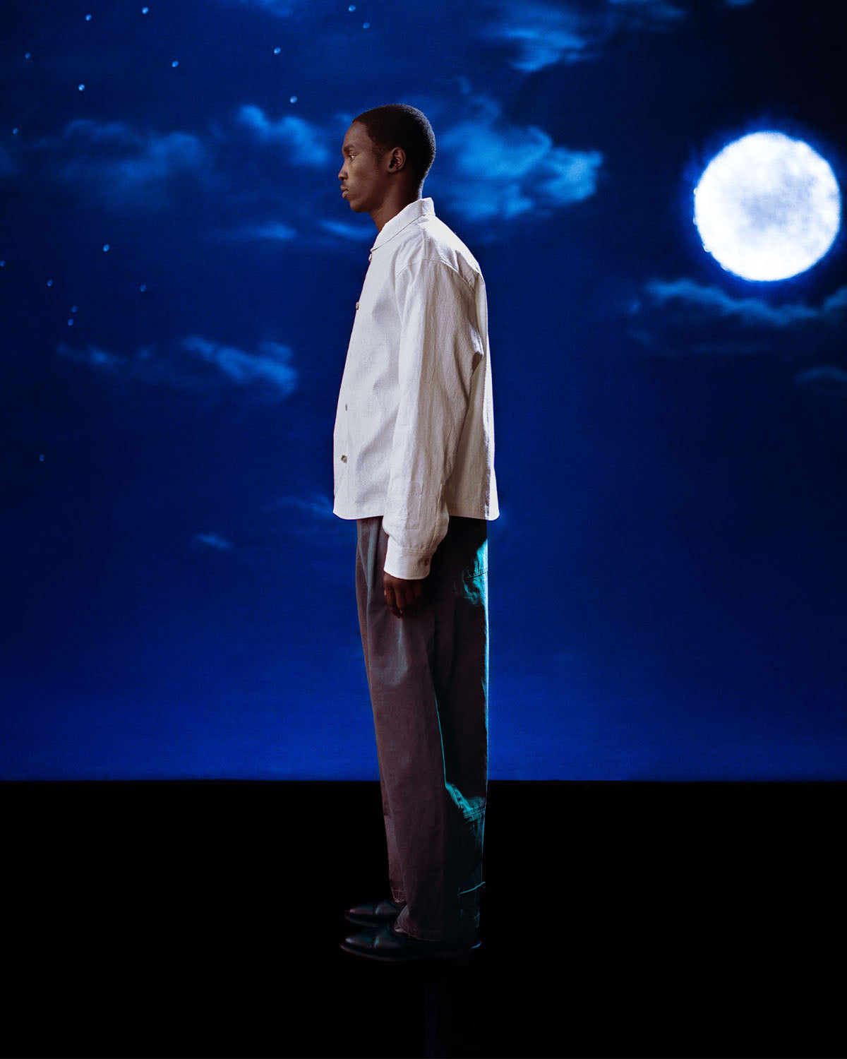 A person stands in profile against a night sky, wearing a white shirt, dark shoes, and Sundae School's CHARCOAL HANBOK CHINO PANTS with a hanbok-inspired silhouette.