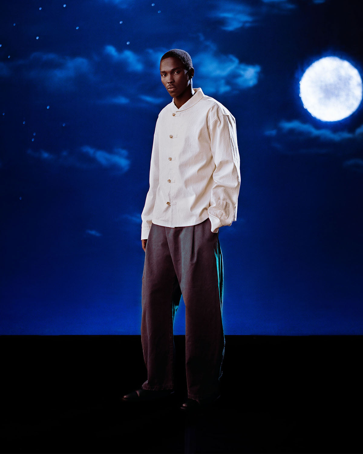 A person stands against a night sky with a full moon and clouds, wearing a white button-up shirt and Sundae School’s CHARCOAL HANBOK CHINO PANTS—hanbok-inspired pants that suit the calm, moody atmosphere perfectly.