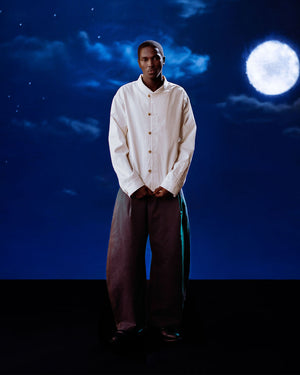 A man stands before a night sky with a full moon and clouds, hands in pockets, wearing a loose white shirt and Sundae School CHARCOAL HANBOK CHINO PANTS, looking directly at the camera.