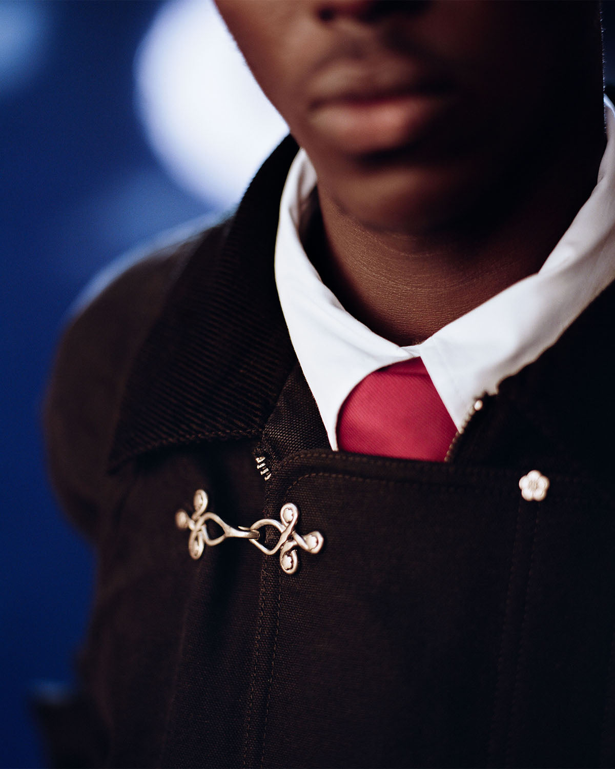 A close-up of a person wearing the [PRE-ORDER] BLACK FIREMAN CLASP JACKET by Sundae School, styled with a white collared shirt and red tie. The blurred background emphasizes the jacket’s unique details.