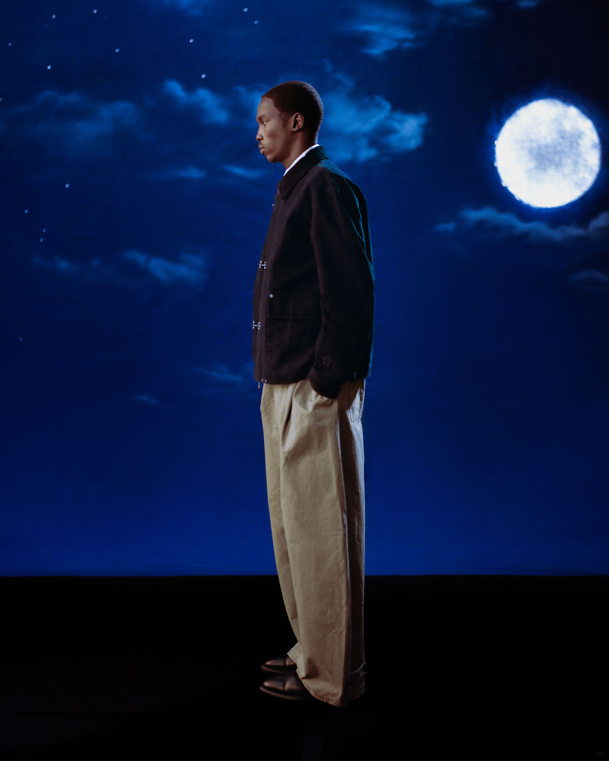 A person stands in profile against a night sky with clouds and a bright full moon, wearing the Sundae School [PRE-ORDER] BLACK FIREMAN CLASP JACKET, light wide-legged pants, and black shoes, hands tucked in their pockets.