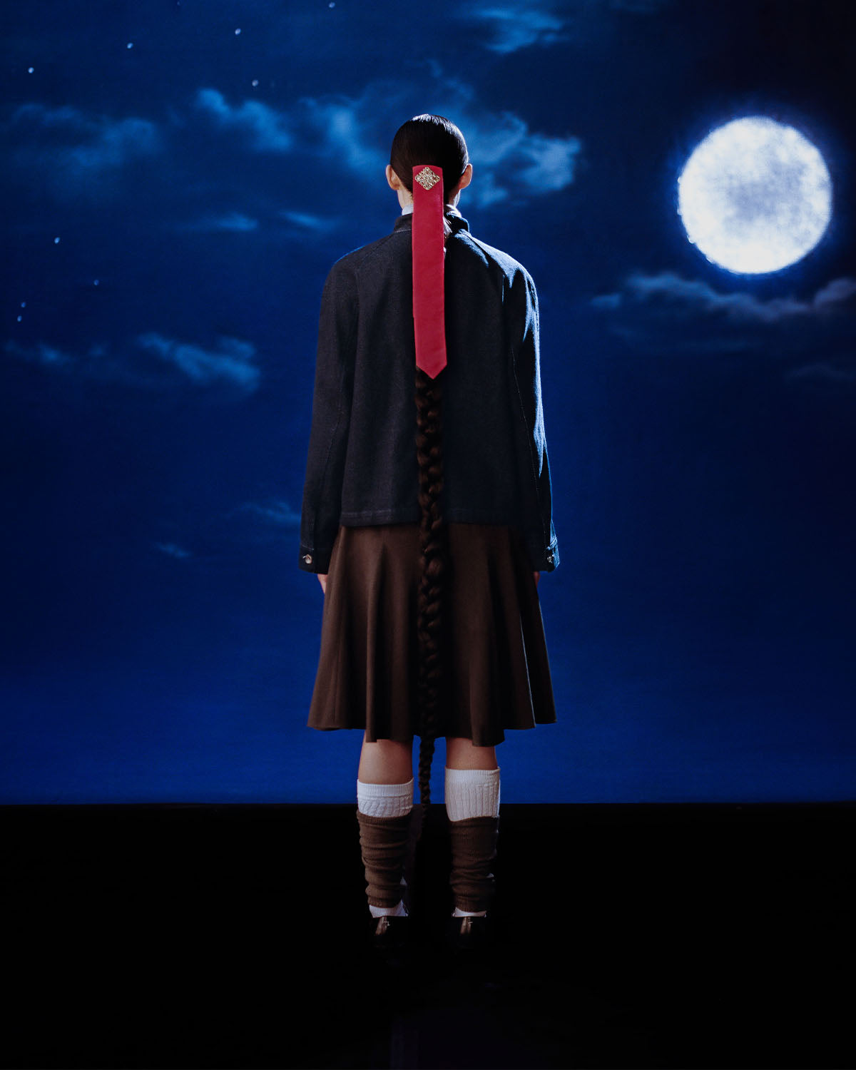 A person with a long braided ponytail tied with a red ribbon faces a full moon, wearing Sundae School's DENIM CHORE JACKET, a brown skirt, tall socks, and brown shoes.