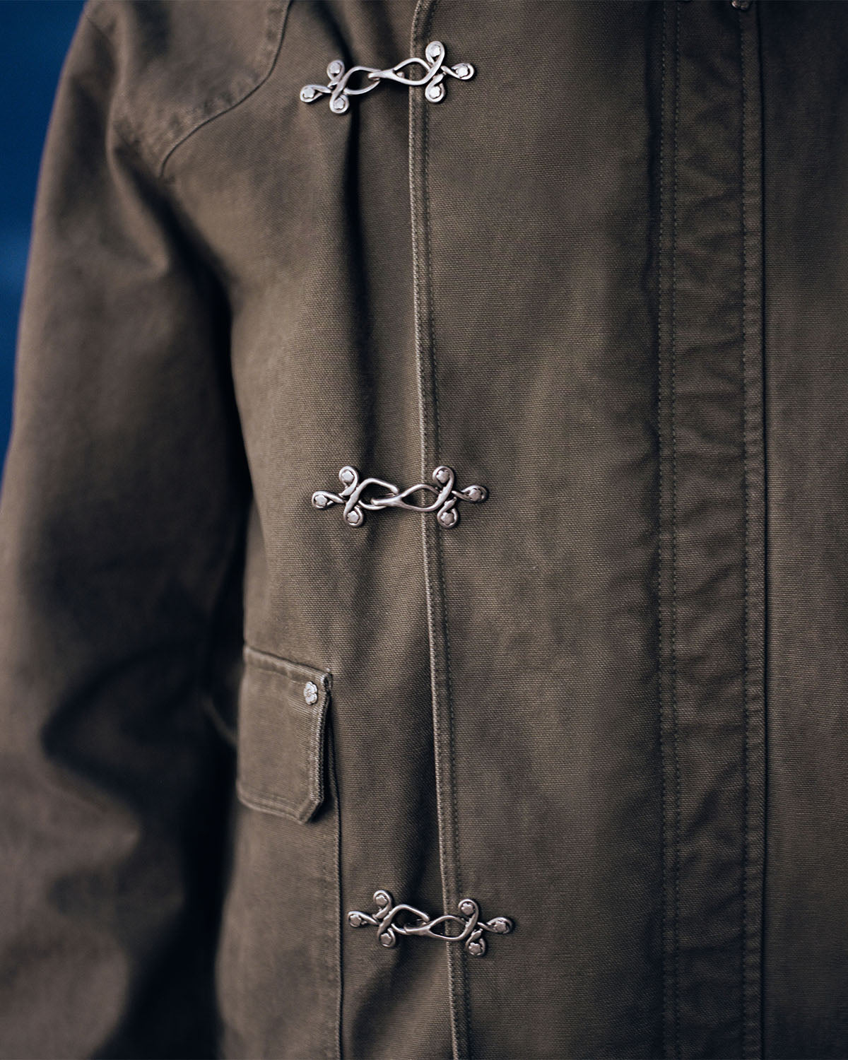 Close-up of Sundae School's [PRE-ORDER] OLIVE FIREMAN CLASP JACKET, a vintage-inspired cotton piece in olive green with silver fireman clasps and a side flap pocket, echoing classic workwear details.