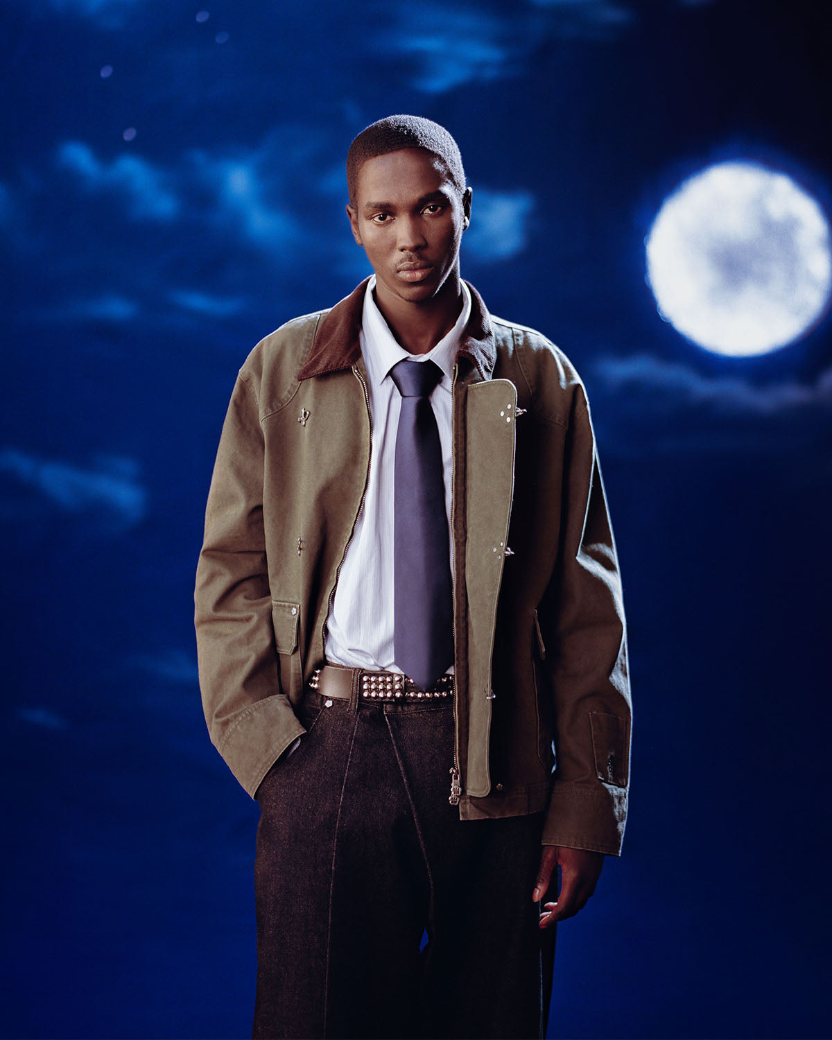 A man stands against a night sky with a full moon, wearing the Sundae School [PRE-ORDER] OLIVE FIREMAN CLASP JACKET over a blue shirt and tie, paired with dark pants. He looks calmly at the camera with one hand in his pocket.