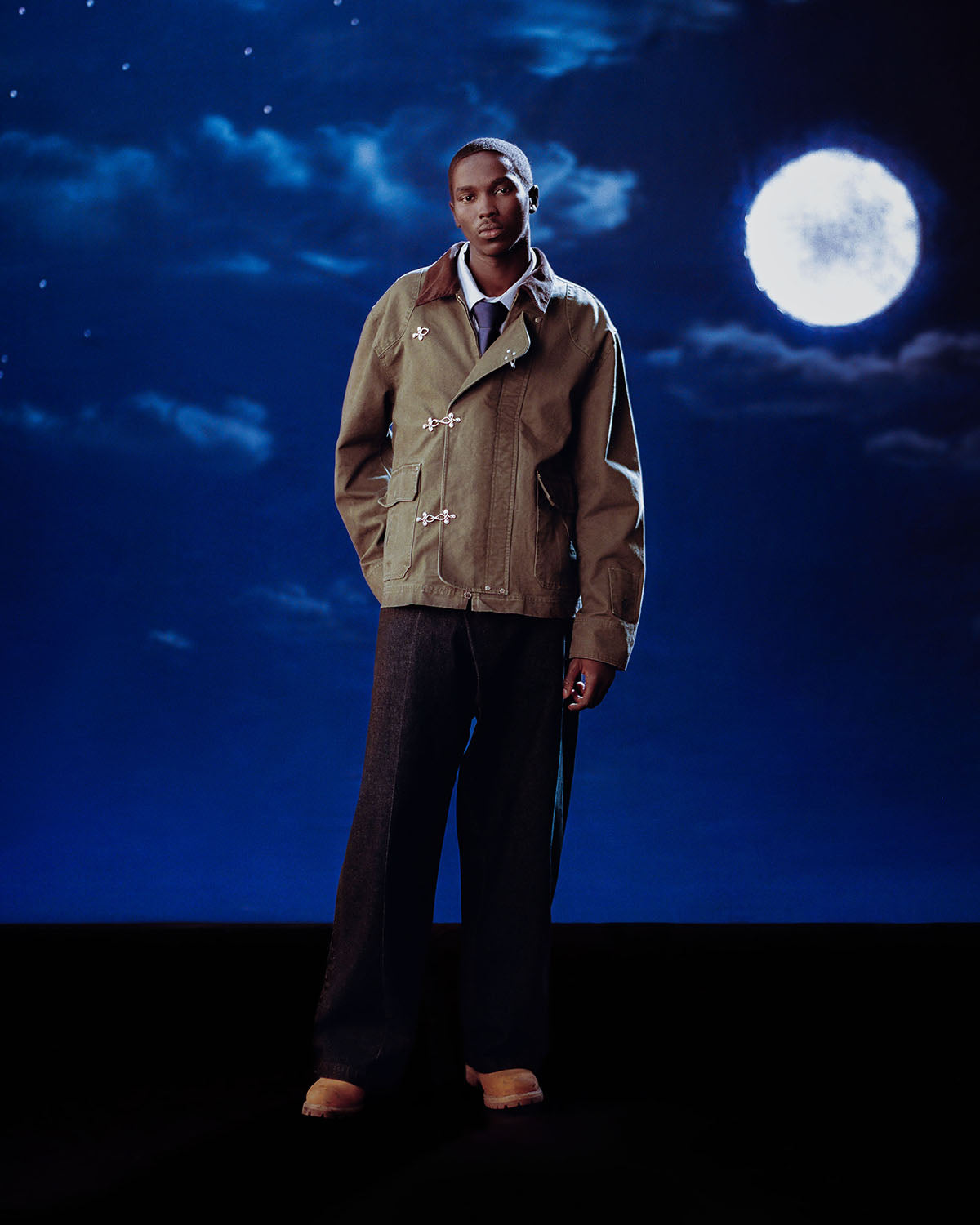 A man stands before a night sky with a full moon, wearing Sundae School's [PRE-ORDER] OLIVE FIREMAN CLASP JACKET, paired with dark wide-leg pants and brown shoes.