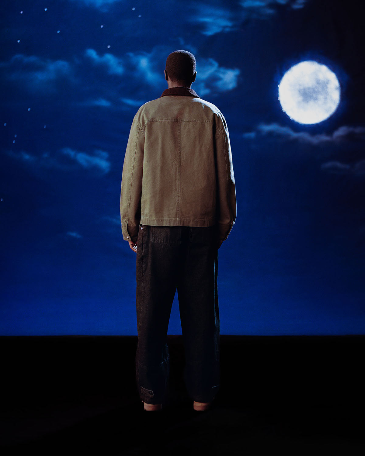 A person faces away from the camera, gazing at a moonlit night sky while wearing Sundae School’s [PRE-ORDER] OLIVE FIREMAN CLASP JACKET, creating a dark and atmospheric mood.