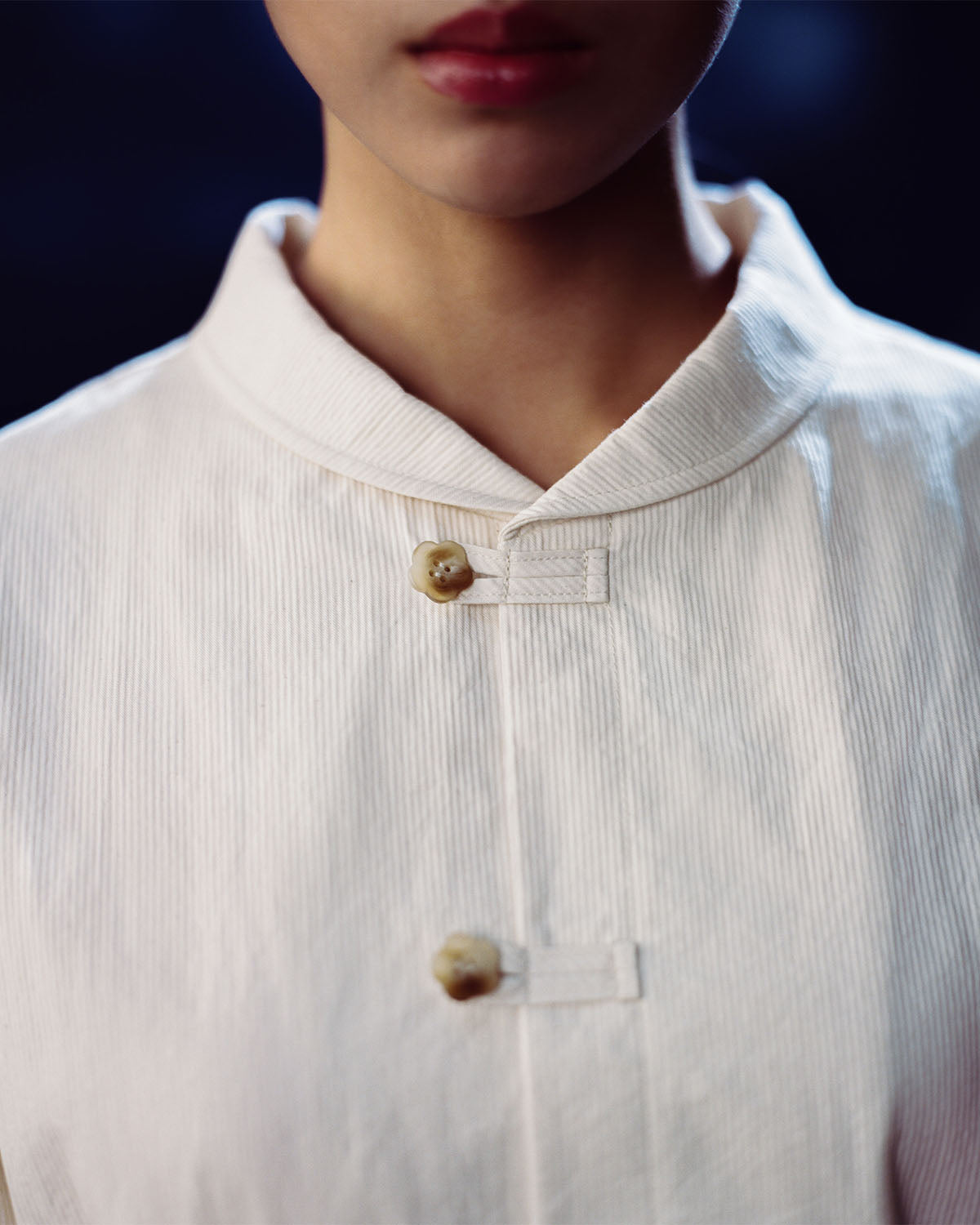 Close-up of a person wearing the IVORY CHINA COLLAR KNOT SHACKET by Sundae School, featuring a relaxed fit, white color, stand-up collar, and two visible buttons. Only the lower face and upper chest are shown against a soft dark blue background.