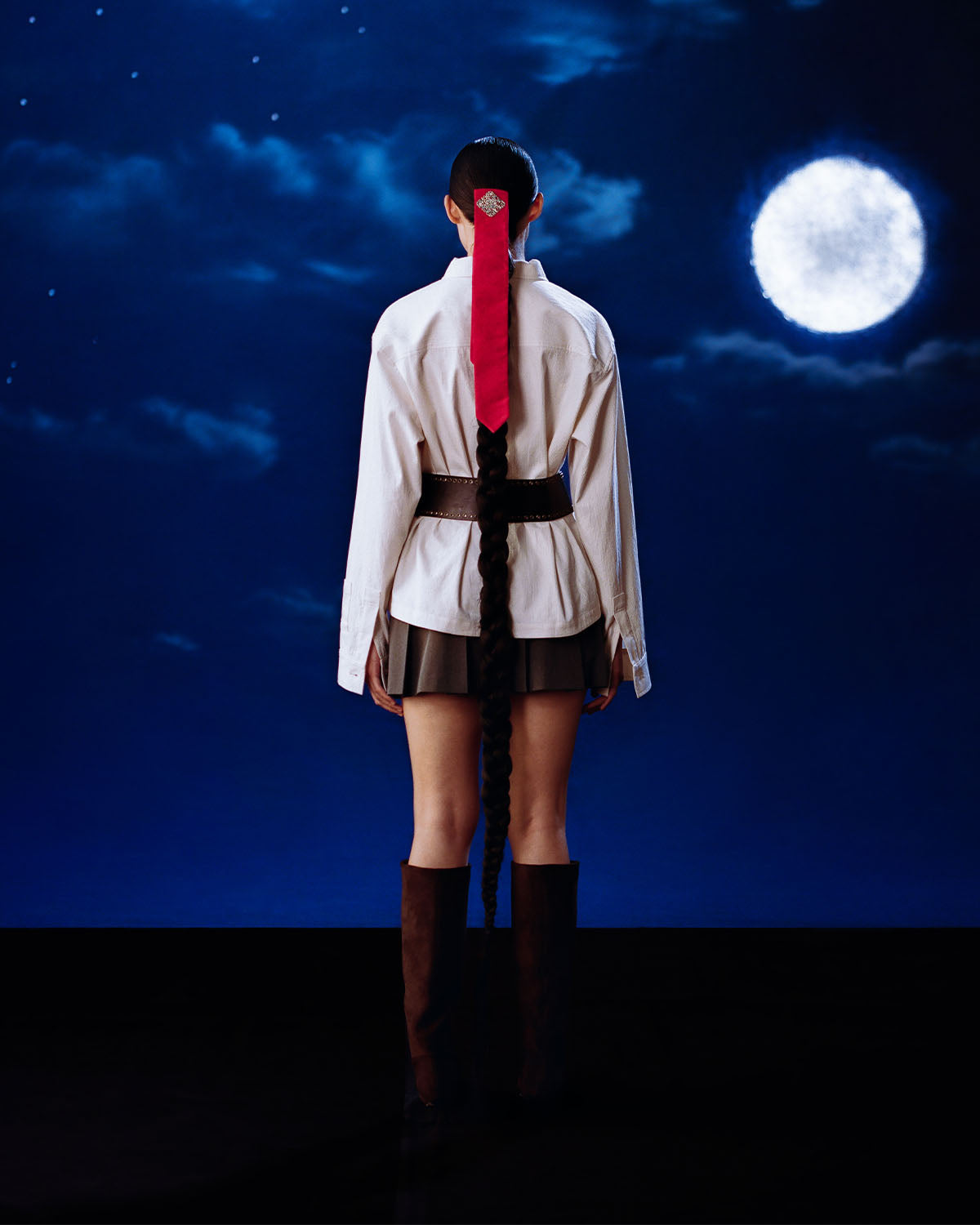 A person with a long braid tied with a red ribbon stands facing a cloudy night sky and full moon, wearing a white shirt, black skirt, wide belt, tall brown boots, and the Sundae School IVORY CHINA COLLAR KNOT SHACKET.