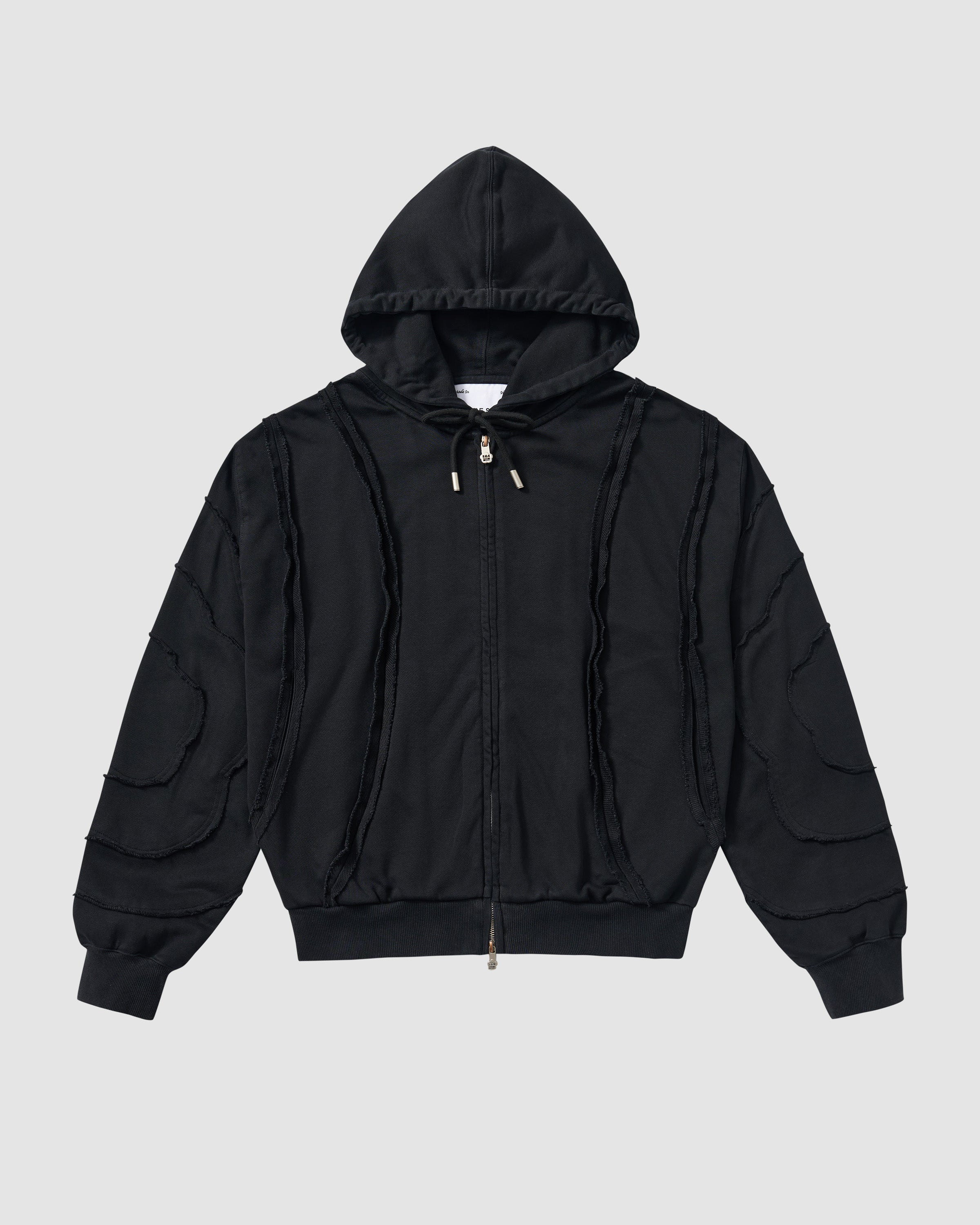 Black Mochi Haze Paneled Hoodie featuring garment washed black cotton with exposed curved seam details throughout body and sleeves, zip-up construction with drawstring hood, ribbed cuffs and hem, relaxed oversized fit silhouette, flat lay product shot on light background.