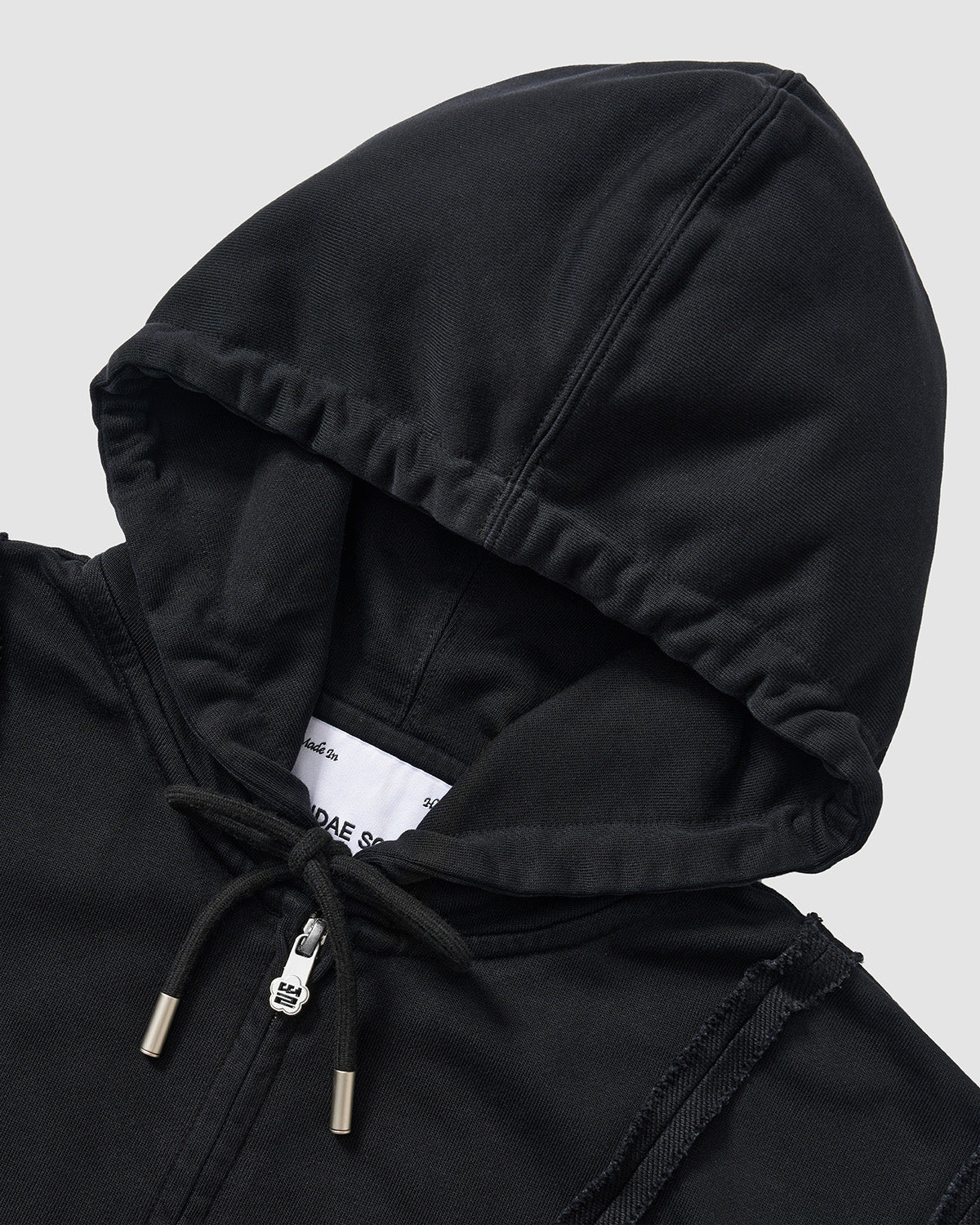Black Mochi Haze Paneled Hoodie close-up detail shot showing drawstring hood with metal aglets, brand label visible inside hood, exposed seam construction details, and garment washed black cotton texture on light background.