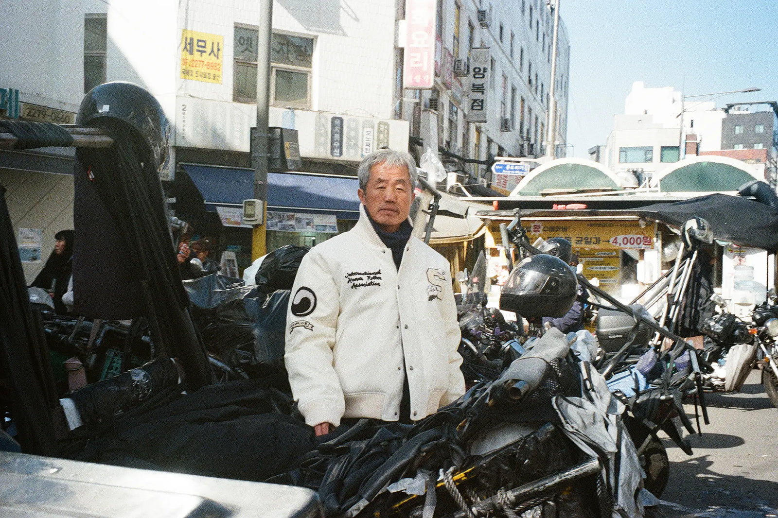 An older man wearing SUNDAE SCHOOL VARSITY JACKET stands in a busy outdoor street market surrounded by parked motorcycles and urban buildings in the background.