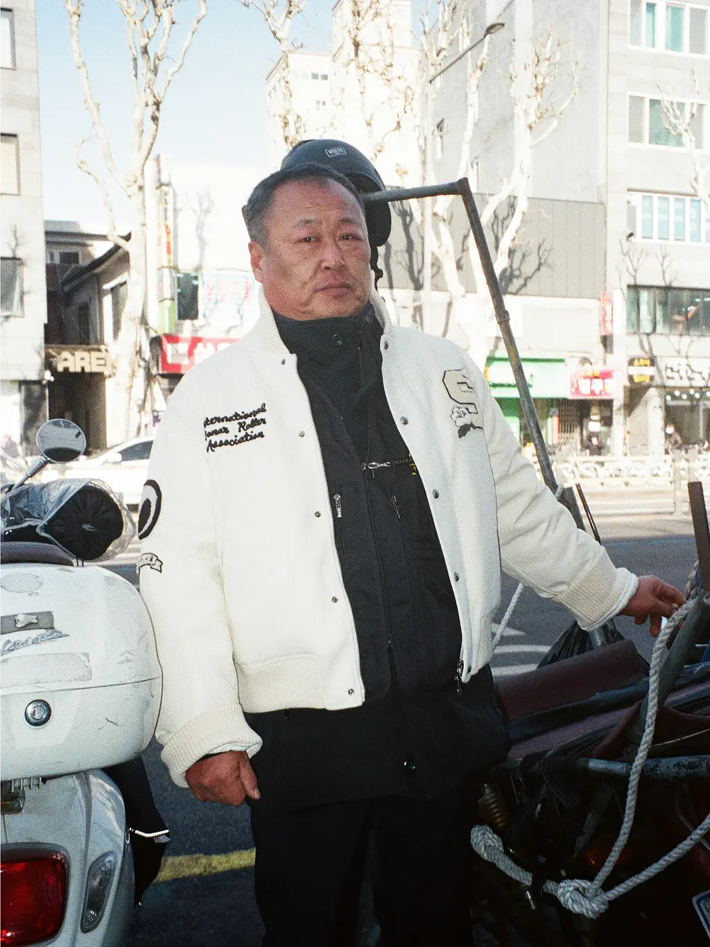 A man wearing SUNDAE SCHOOL VARSITY JACKET over a black shirt and pants, standing outdoors next to a white scooter in an urban street setting.