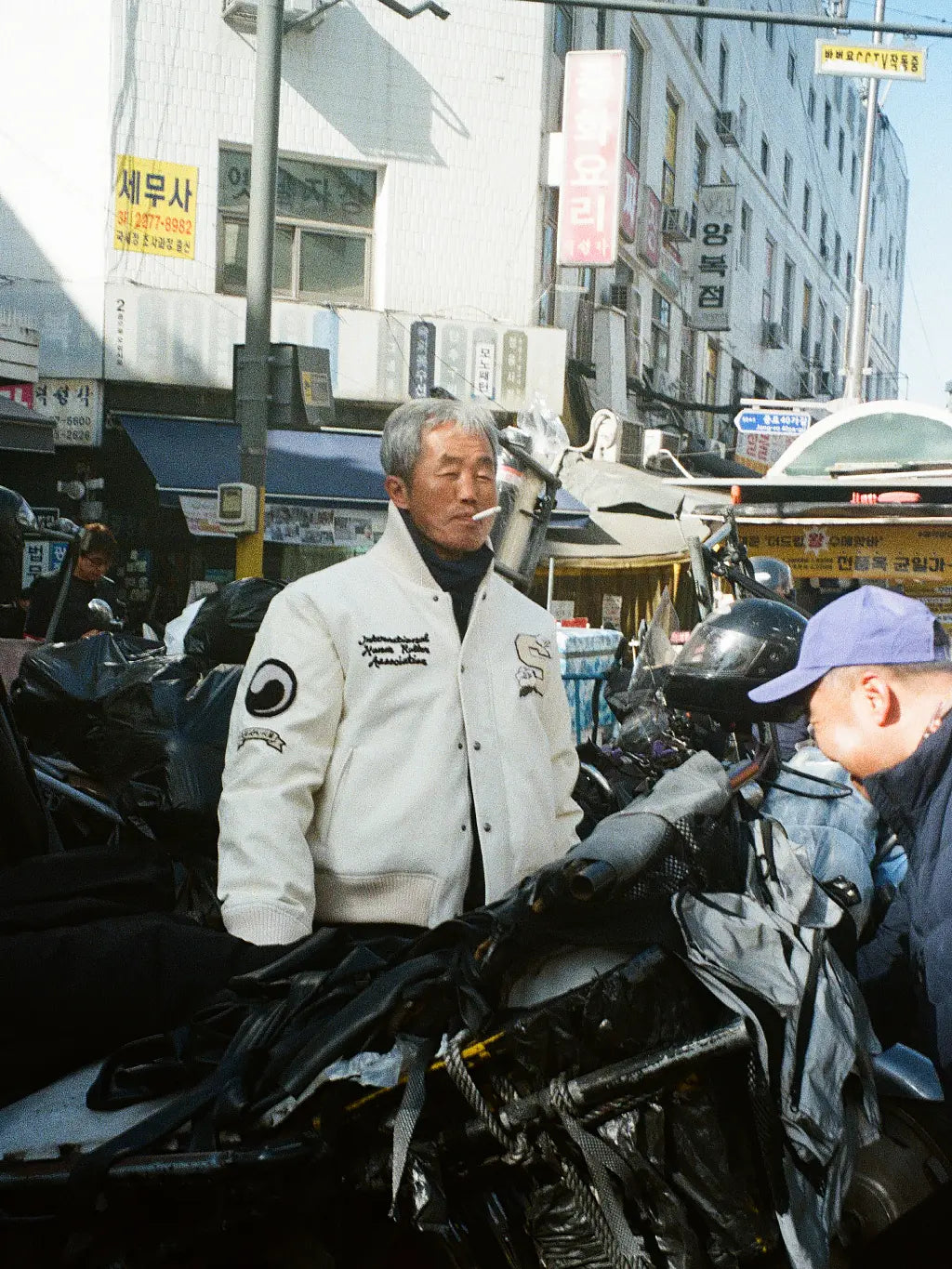 A man in SUNDAE SCHOOL VARSITY JACKET standing on a street behind a motorcycle with a cigarette in his mouth, with buildings and Korean signs in the background.