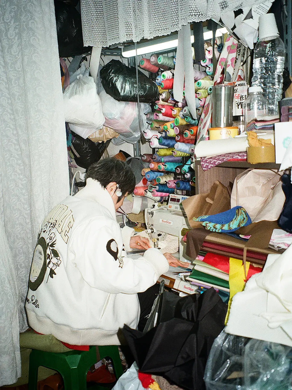An old Korean lady  wearing SUNDAE SCHOOL VARSITY JACKET sits at a sewing machine inside a cluttered, busy workshop filled with fabrics, spools of thread, and sewing supplies.