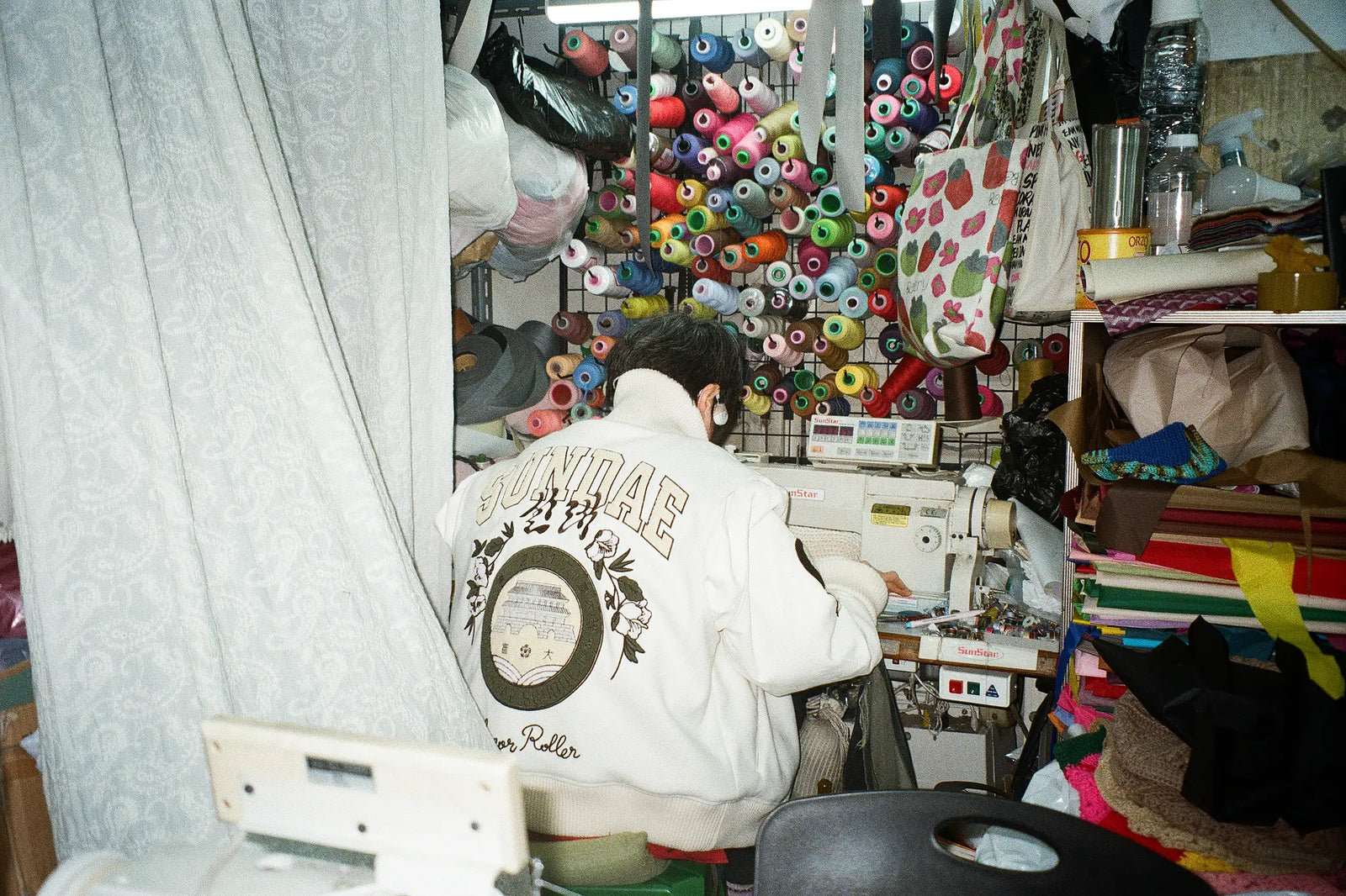 An old Korean lady seen from behind wearing SUNDAE SCHOOL VARSITY JACKET, seated at a sewing machine in a cluttered sewing room filled with colorful thread spools.