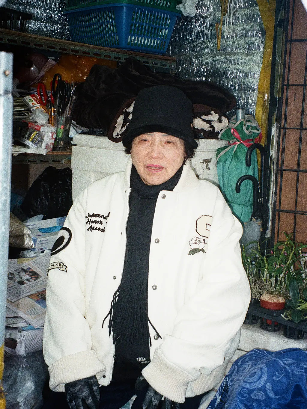 An old Korean lady wearing SUNDAE SCHOOL VARSITY JACKET, a black scarf, and a black hat, standing in a cluttered indoor storage area with shelves, plastic bins, and household items.