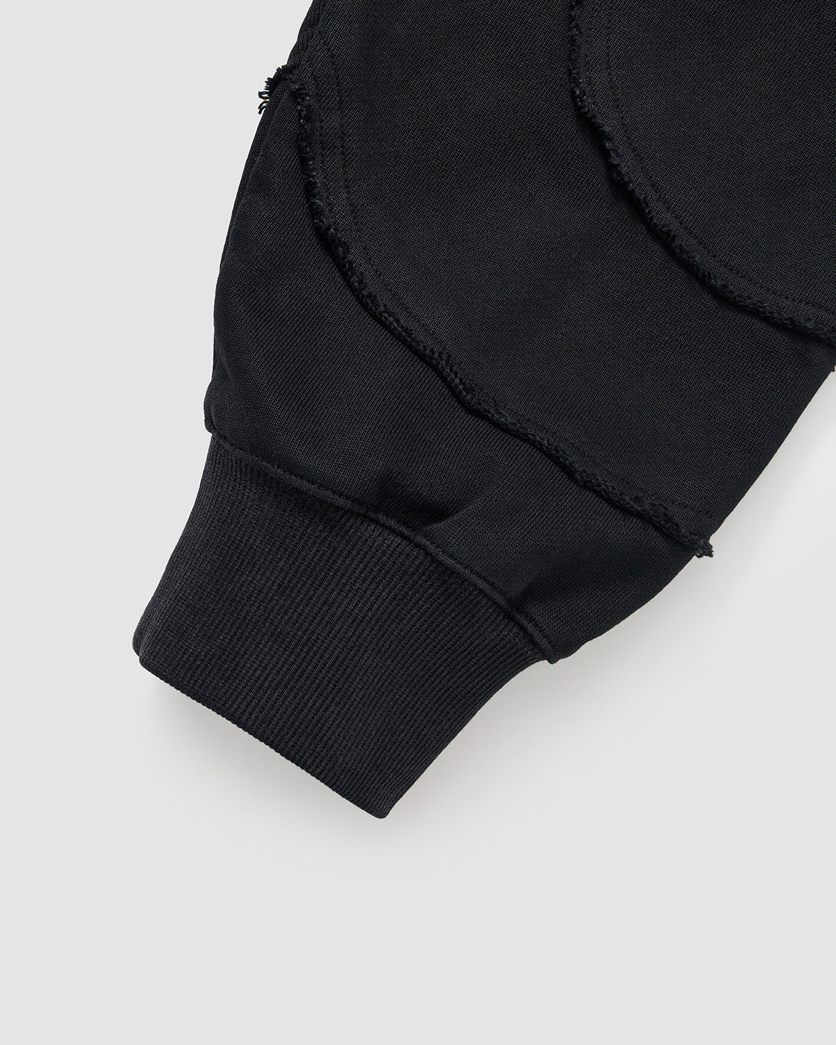 Black Mochi Haze Paneled Hoodie sleeve cuff detail showing exposed curved seam construction on black garment washed cotton fabric, ribbed cuff band, and distressed finishing details, flat lay on light background.