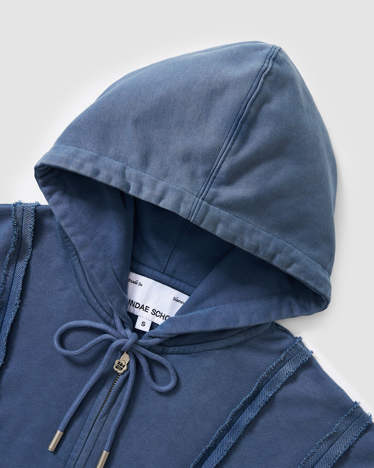 Indigo Mochi Haze Paneled Hoodie hood detail showing drawstring ties with metal aglets and brand label, exposed curved seam construction visible, garment washed indigo cotton texture, flat lay on light background.