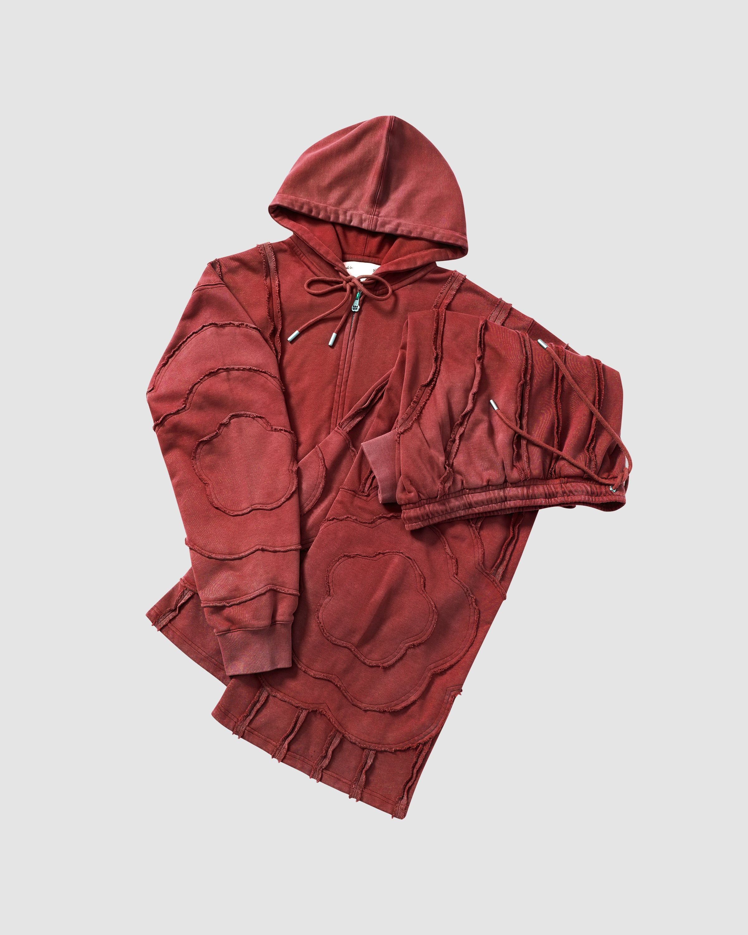 Red Mochi Haze Paneled Hoodie and Sweatpants flat lay detail shot showing washed red cotton texture, exposed curved seam construction, and fabric draping, flat lay on light background.