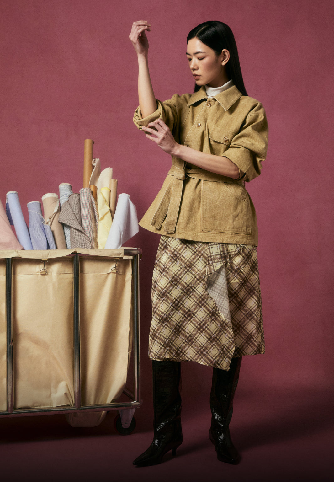 A model wearing a tan Asymmetric Work Jacket cinched with a belt and a plaid skirt, standing next to a vintage fabric cart filled with colorful textile rolls against a textured magenta background.