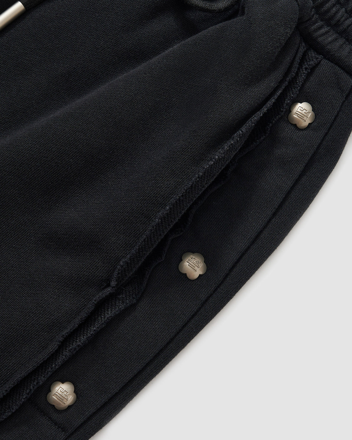 Black Fade Black Snap Sweatpants close-up detail showing custom dome snaps with branded hardware along side seam, vintage fade washed black cotton texture with raw edge finishing, flat lay on light background.
