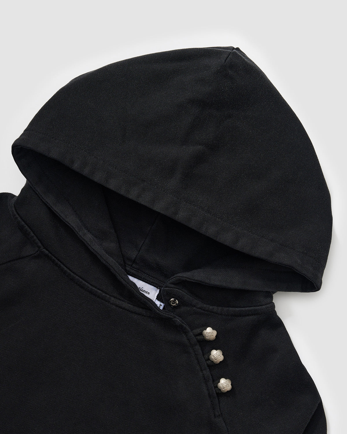 Black Fade Black Hanbok Hoodie hood construction detail showing asymmetric closure with custom dome snap hardware and brand label, garment washed black cotton fabric, flat lay on light background.