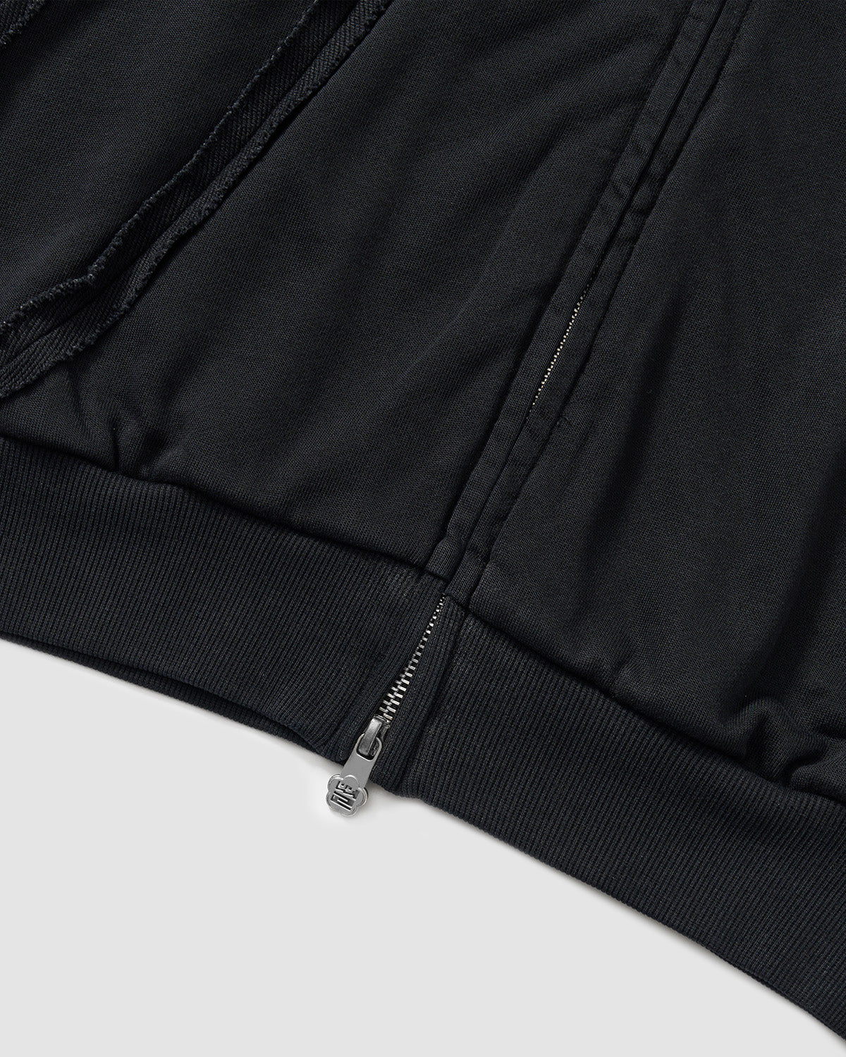 Black Mochi Haze Paneled Hoodie hem detail showing zip closure with branded hardware, ribbed waistband, exposed seam construction, and garment washed black cotton texture, flat lay on light background.
