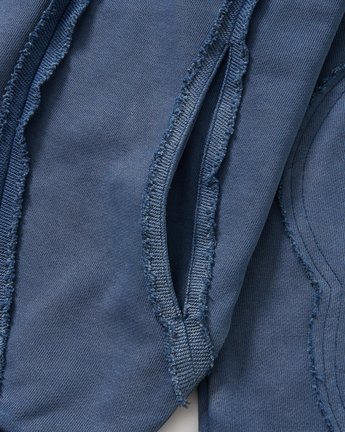 Indigo Mochi Haze Paneled Hoodie close-up detail showing exposed curved seam construction with frayed edges on garment washed indigo cotton fabric, flat lay on light background.