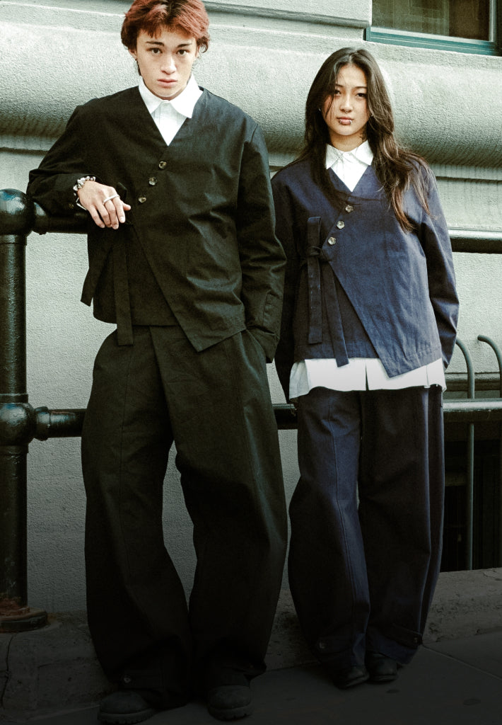 One asian boy and one asian girl standing leaning against a metal fence wearing Sundae School Hanbok Blazer and Hanbok Balloon pants in different colors, one in navy and one in black.