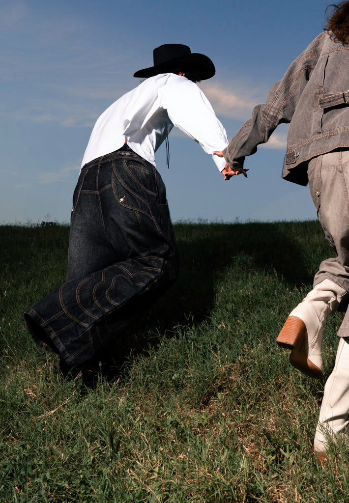 Two people running in the fields holding hands wearing the Black Mochi Spiral Denim and Black Mochi Paneled Jorts from a collaboration collection between Sundae School and Jooniverse.