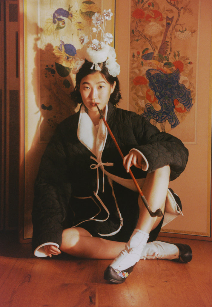A girl wearing the black side of Black Ivory Reversible Flower Quited Jeogori Fleece biting gonbangdae, Korean traditional smoking pipe, sitting on the Korean traditional room divider.