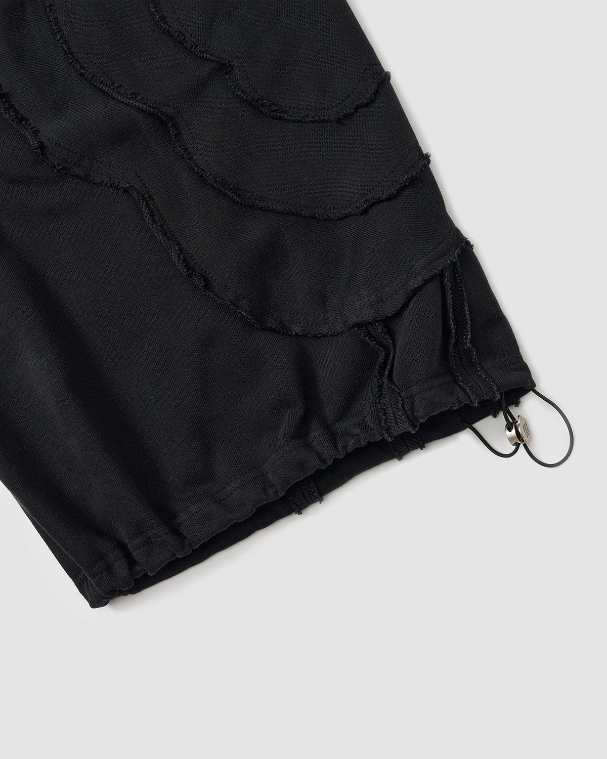 Black Mochi Haze Paneled Sweatpants hem detail showing curved exposed seam construction with frayed edges, drawstring closure at ankle, and distressed finishing on garment washed black cotton, flat lay on light background.