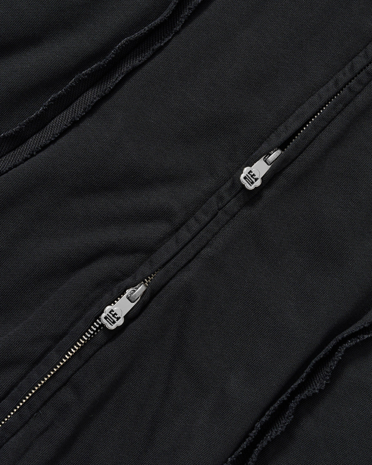 Black Mochi Haze Paneled Hoodie close-up detail shot showing dual zip pulls with branded hardware on washed black cotton, exposed seam construction visible, flat lay on light background.