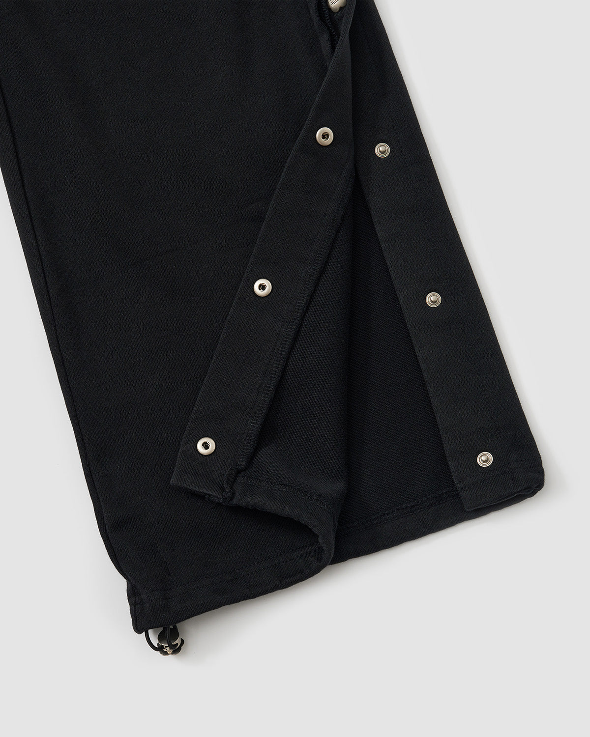 Black Fade Black Snap Sweatpants hem detail showing custom dome snaps extending to ankle with raw edge finishing, vintage fade washed black cotton fabric, flat lay on light background.