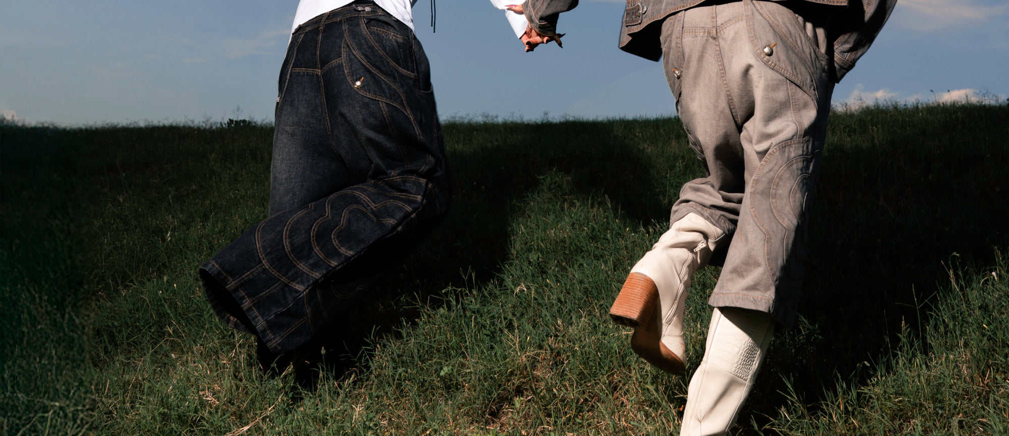 Two people running in the fields holding hands wearing the Black Mochi Spiral Denim and Black Mochi Paneled Jorts from a collaboration collection between Sundae School and Jooniverse.