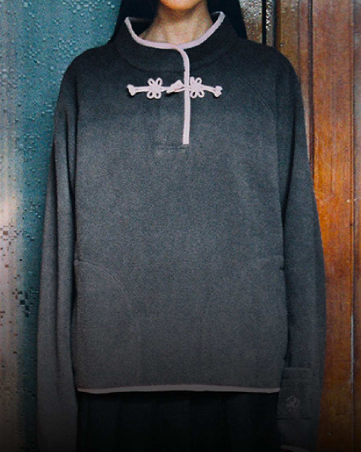 In front of wooden doors indoors, a person looks directly at the camera, wearing Sundae School’s CHARCOAL BUTTERFLY FLEECE PULLOVER—a gray mock-neck polar fleece with tteokbokki toggles—paired with a gray hat, black pleated skirt, white socks, and black shoes.